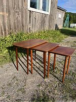 Three teak nesting tables arranged side by side outdoors on gravel in front of a wooden wall