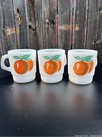 Front view of three white milk glass mugs with orange fruit and green leaf graphics, arranged side by side on a black surface, with a wooden fence background.
