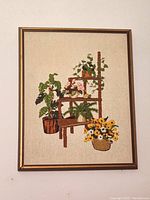 Full view of framed vintage crewel embroidery artwork featuring a wooden plant stand with multiple potted houseplants, including a plant in a basket with yellow and white flowers.