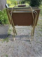 Set of four folded vintage TV trays standing upright and nested on the rolling cart outdoors on a gravel surface, showing the back side of the trays and the cart.