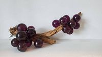 Single photo showing a large cluster of purple Lucite grapes attached to a realistic wood-textured stem, photographed against a plain background.