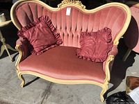 Front view of the Louis XVI style love seat showing pink velvet upholstery, cameo shaped back with vertical channel tufting, cream carved wood frame, and two ruffled cushions.