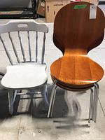 Photo showing one white painted wooden kitchen chair with turned legs and spindle backrest alongside one veneer wood stacking chair with metal legs.
