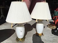 Two white ceramic table lamps with brass or gold-tone metal bases and white pleated shades.