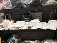 Shelving unit with neatly folded white and off-white sheets and linens, pillows behind them, and other linen items stored in plastic bags on top shelf.