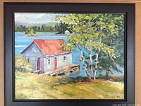 Framed oil painting showing lakeside scene with gray boathouse with red roof and doors, trees, water, and distant hills.