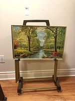 Photo showing a metal TV tray with a lush green forest and river landscape. The tray has a brown handle and gold edges and is held on a metal stand with wheels.