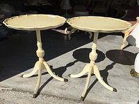 Two round wooden side tables with scalloped edges and claw feet standing outdoors on concrete floor with shadows.