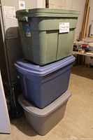 Three large rectangular plastic storage bins stacked. Top bin is green, middle blue, bottom gray. Each bin has a lid. The bins are situated in a garage environment.