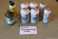 Photo showing lot of 7 cans on beige surface, including Super Tech R-134A refrigerant and PAG oil cans.