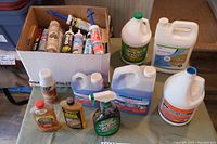 Photo of a cardboard box with multiple tubes of caulk and sealant with caps, positioned beside various large bottles of cleaning products including Simple Green and Rust & Forget jugs.