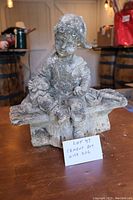 Front angle showing the entire cement statue of a boy wearing a baseball cap seated on a bench with a small dog on his lap, displaying signs of weathering and dirt accumulation.