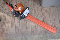 Full view of gas hedge trimmer with orange blade guard laying on wooden floor.