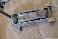 Close-up of the gray metal floor jack on wooden floor, showing hydraulic lift and lifting pad with dirt and wear.