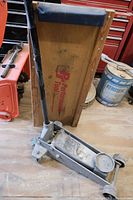 Floor jack leaning against a wooden mechanic creeper on a workshop floor with surrounding tools and equipment.