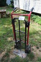 Photo of a red and black heavy duty hand truck with pneumatic tires and foldable metal toe plate, shown outdoors on grass.