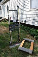 Photo shows a metal hand truck with two wheels and handles alongside a rectangular furniture dolly with a wooden frame and carpeted surface. The items are positioned outdoors on grass next to a white sided building.