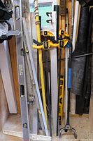 Full view of assorted tools including Stanley Stanley FatMax levels, pole saw with rope, clamps, and metal squares arranged upright in a workshop corner.