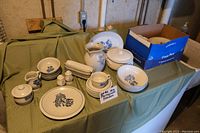 Wide shot showing all Pfaltzgraff dishware items arranged on table with green cloth background.