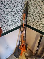 Full view of several roofing tools standing against corner wall, includes shovels and rakes with orange handles and black metal parts.