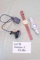 Photo of two wristwatches and Fitbit Charge 5 fitness tracker with charger and power adapter on a green patterned surface and a handwritten label reading 'Lot 98 Watches & Fit Bit'.