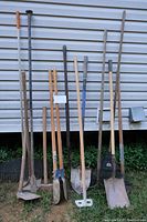 Photo showing ten yard tools including rakes, hoes, shovels, and forks arranged vertically against house siding.