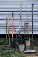 Full lot of assorted yard tools leaning against a building, showing wood and metal tool handles including rakes, shovels, a fork, and a push broom.