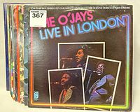 Photo showing stack of 13 vinyl record albums, front album is Live In London by The O'Jays with visible wear.