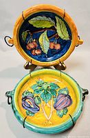 Two vintage Italian pottery plates displayed on metal stands, showing colorful hand-painted fruit and leaf designs, both with attached metal wall hanging hooks.