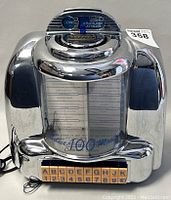 Front view of Crosley novelty jukebox cassette player showing chrome reflective surface, coin slot, buttons A-K with numbers, and song list