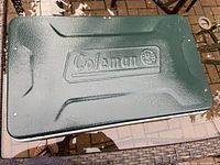 Closed green Coleman camping stove lid with embossed brand logo.