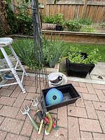 Overview photo showing garden hand tools, plant support, black seedling trays, white bucket and blue hose reel on patio with garden beds in background