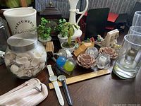 Wide angle photo of assorted knick knacks including seashell jar, small plants, ceramic candle holders, glass spheres, watches, and others.