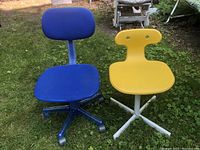 Two small desk chairs placed outdoors on grass: a blue fabric swivel chair with wheels and a yellow plastic swivel chair without wheels.