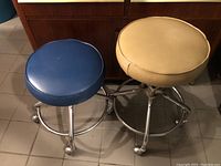 Top view showing one blue and one beige round padded stools on chrome wheel bases.