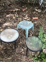 Three round garden tables in outdoor garden setting on dirt ground with dry leaves and plants around. One table blue plastic, two with black metal legs and glass tops.