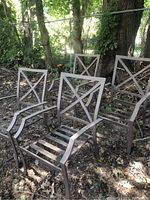 Four grey metal outdoor lawn chairs arranged on an outdoor ground covered with leaves, showing crossed backrest and slatted seat design.