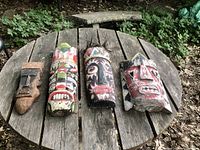 Four wooden tiki masks displayed on a wooden outdoor table surrounded by leaves and dirt ground, showing weathered paint and carved details.