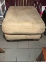 Front view of beige faux suede ottoman showing rectangular tufted cushion and texture of upholstery.