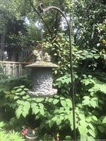 Full view of grey ceramic bird feeder hanging from curved metal garden stake, surrounded by green foliage.