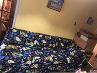 Three-seat couch covered with a tropical themed slip cover that shows colorful tropical leaves, flamingos, monkeys and other wildlife on a dark background, against a light yellow wall.