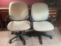 Two grey office chairs side by side on tiled floor against a wooden desk. Both chairs have black armrests and five caster wheels, with light grey fabric upholstery showing some wear.