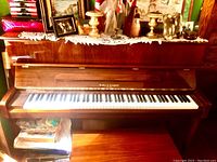 Full view of Kohler & Campbell upright piano with keyboard revealed, cherry polish finish, various items on top, bench in lower part of image.