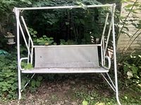 Full view of the metal frame backyard swing showing the entire swing structure, mesh seat and backrest, and support frame with rust and surrounding vegetation.