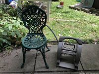 Green metal Victorian style garden chair next to portable hose reel with green grass and plants in background.