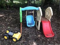 Photo showing plastic toddler slide and swing set next to yellow and black toy truck outdoors on ground covered with leaves.