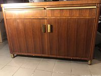 Front view of the vintage wood cabinet showing brass handles and legs, wood chip on bottom front edge.