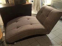 Side angle view of taupe chaise lounge showing curved seat and tufted fabric upholstery with some visible wear.