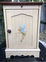Front view of pale yellow wood cabinet with Tinkerbell decal on door, showing round knob and decorative bottom trim.