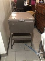 Front view of taupe vintage metal file cabinet with wheels, handle, and lower shelf showing wear and scratches.
